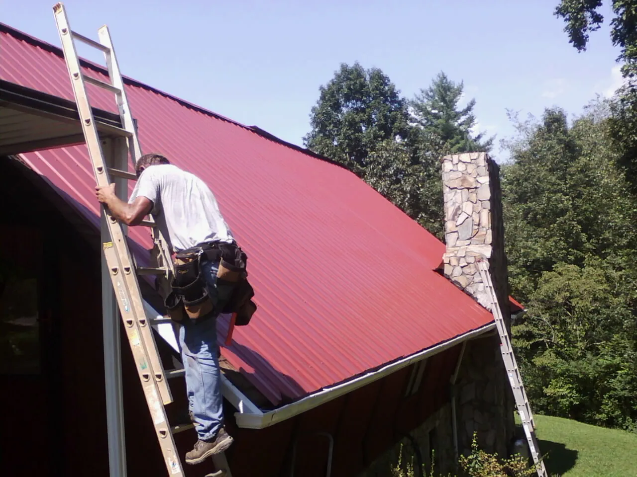 Roofer on a ladder beside a red metal roof and stone chimney — Soffit & Fascia Repair by Roofing Brothers in Mountain Home Afb